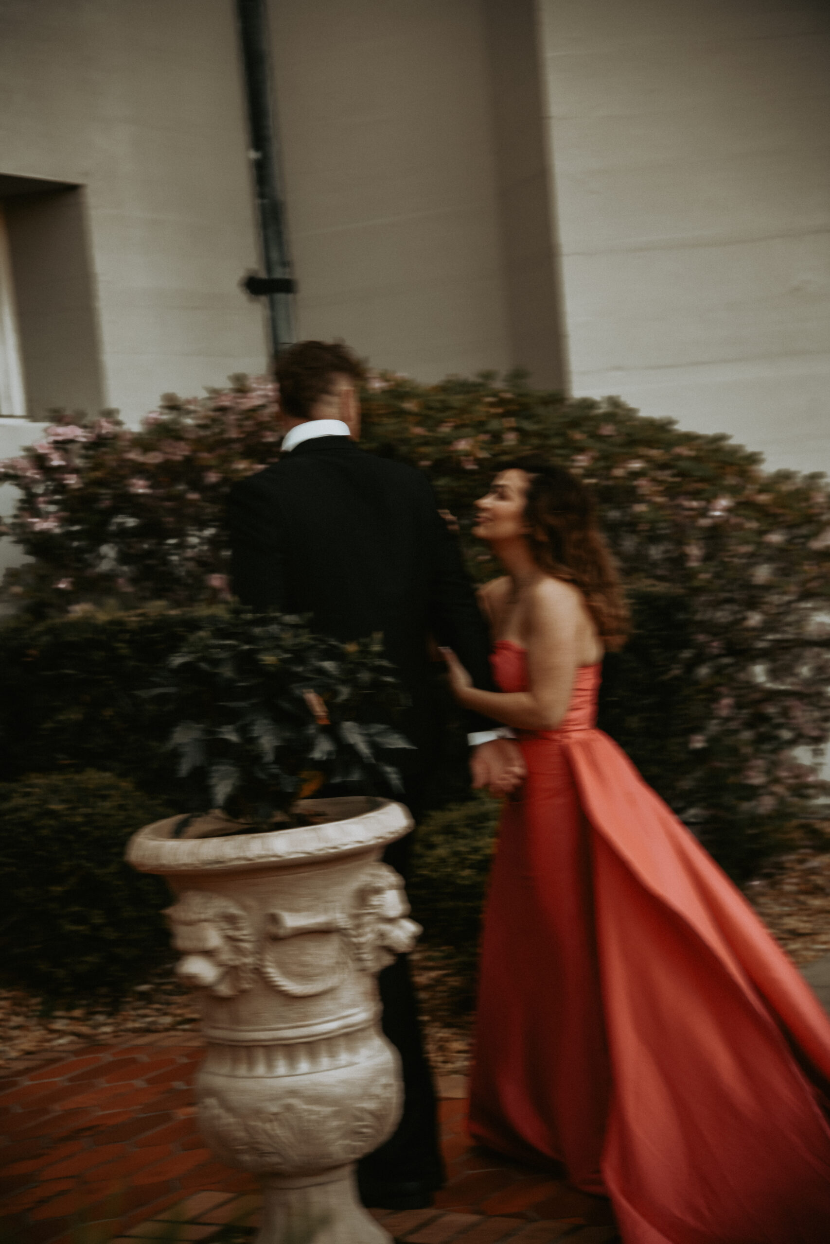 A couple in evening wear, she in a flowing coral gown, walking hand in hand past a stone garden urn in soft cinematic motion blur