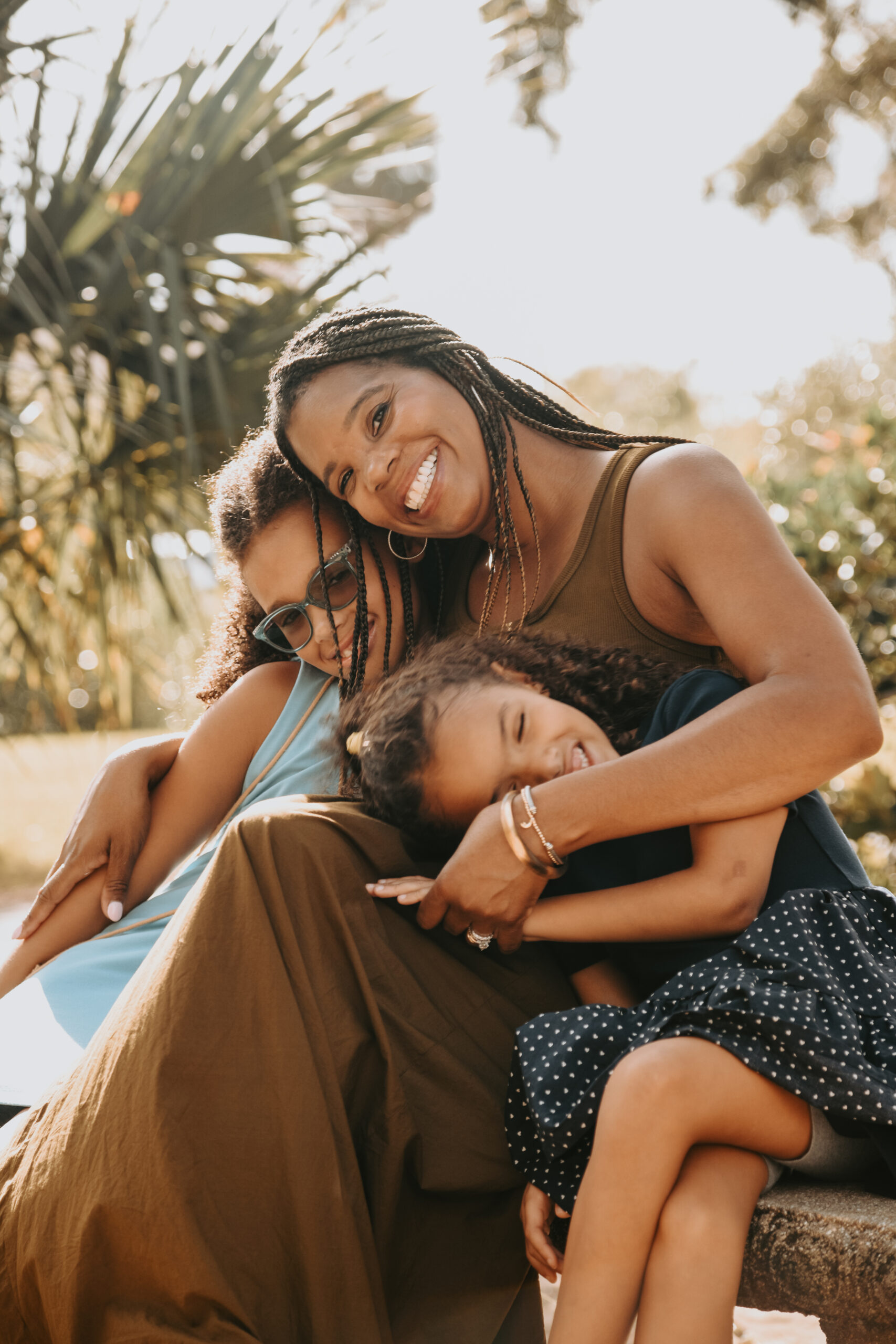 A mother laughing with her two daughters wrapped in her arms on a stone bench beneath palms in golden evening light