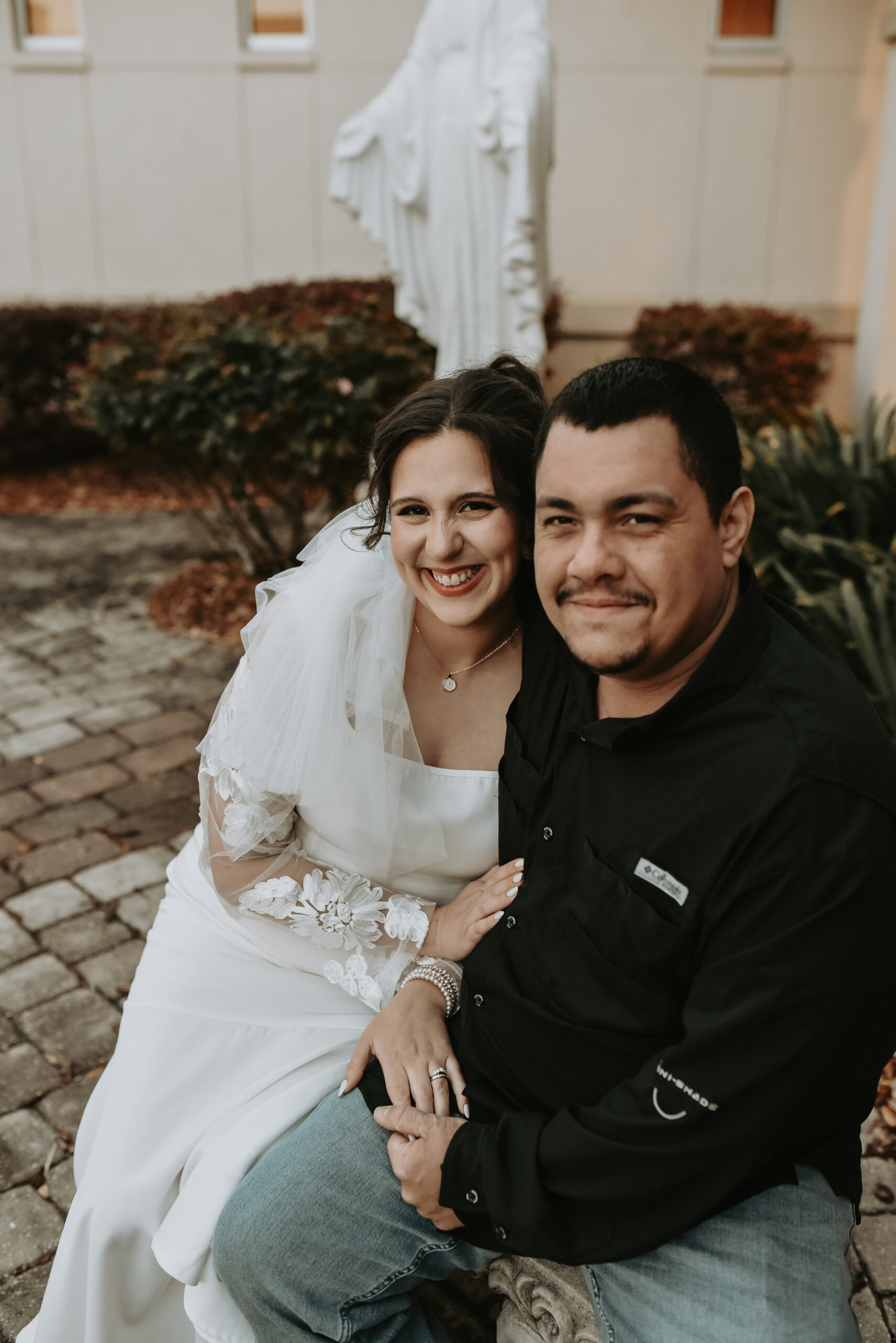A bride in her veil and her groom sitting close together in a courtyard, both smiling warmly