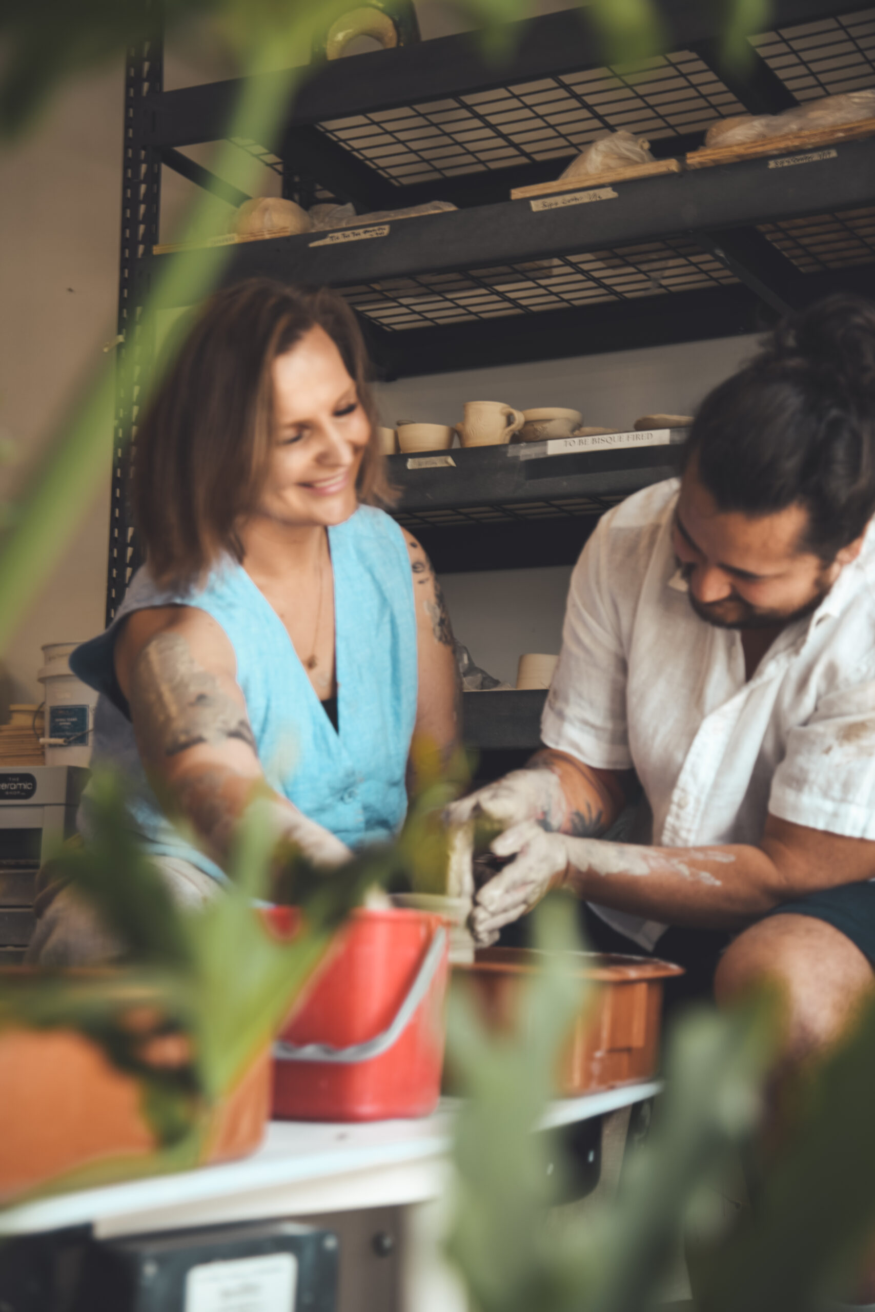 A couple sharing a pottery wheel in a sunlit studio, photographed candidly through green leaves