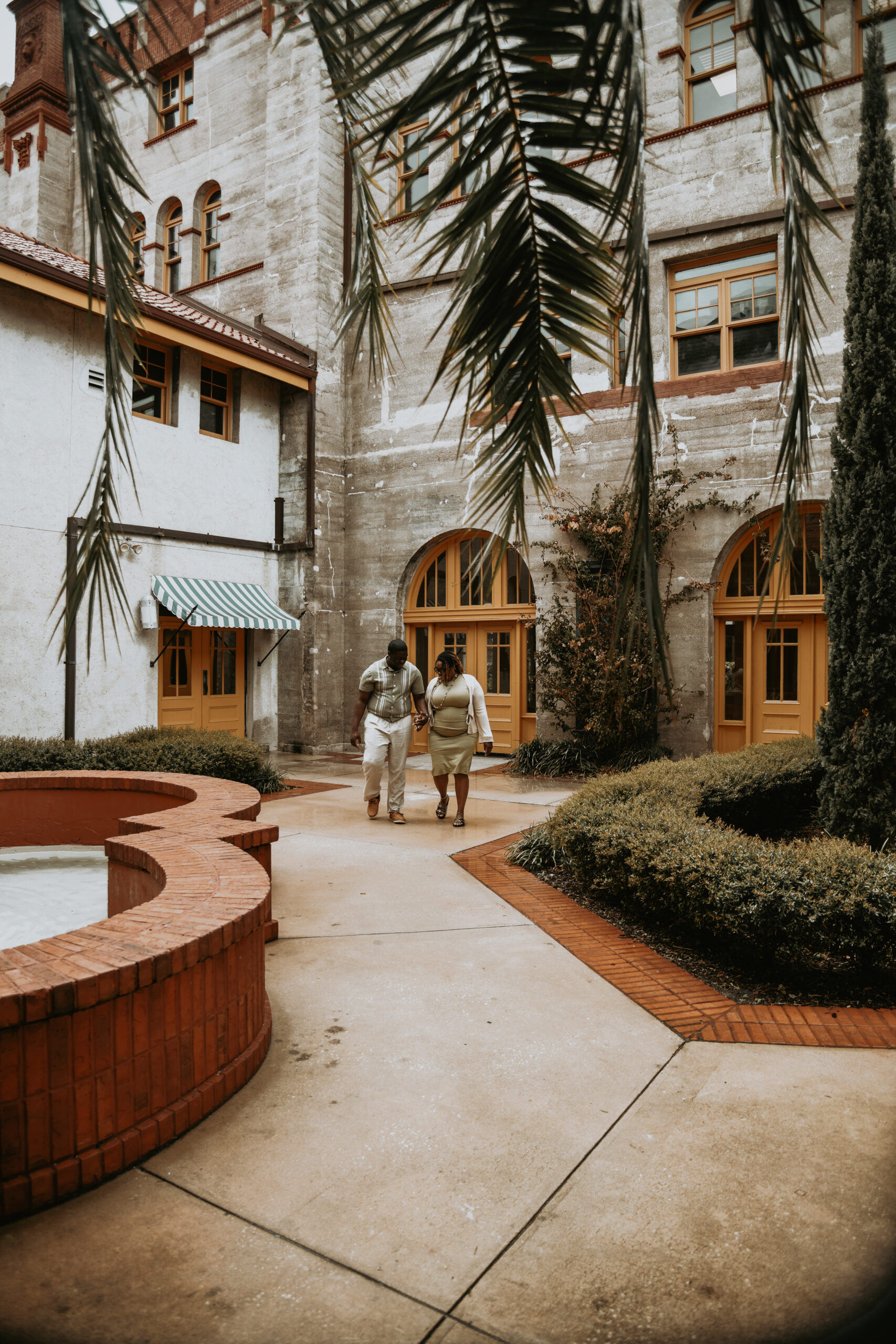 A couple strolling hand in hand through a palm shaded courtyard past a brick fountain in the old city